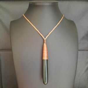 May include: A long, dark green pendant necklace with a copper-colored wrapped top and a braided tan cord. The pendant is displayed on a black mannequin, showcasing the necklace's design and color contrast.
