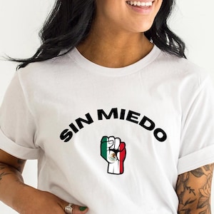 May include: White t-shirt with the text "SIN MIEDO" in black lettering in an arc above a raised fist with the Mexican flag colors.