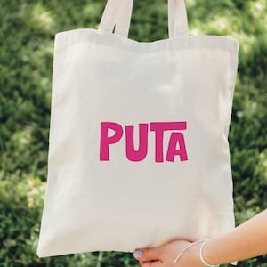 May include: Cream-colored tote bag with the word "PUTA" printed in bright pink. The bag is held by a person against a backdrop of green foliage. The tote bag has two handles.