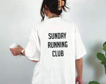 May include: White t-shirt with the text "SUNDAY RUNNING CLUB" in black block letters. The shirt is worn by a person holding a white coffee cup. A watch is visible on the person's wrist.