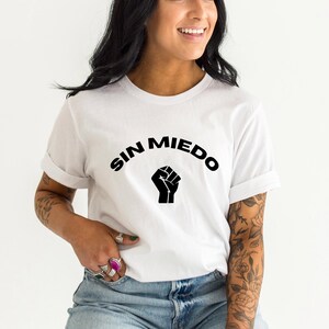May include: White t-shirt with black text that reads "SIN MIEDO" in a semi-circle with a raised fist in the center.