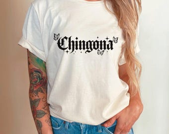 May include: Cream-colored t-shirt with the word "Chingona" in a gothic font, adorned with butterfly and star graphics. The shirt is paired with blue jeans.
