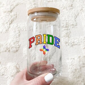May include: A clear glass reusable can with a wooden lid and a rainbow colored straw. The can has a rainbow colored "PRIDE" graphic with a rainbow colored symbol below it.