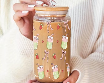 Puede incluir: Un vaso reutilizable de vidrio transparente con tapa de bambú y una pajita. El vaso tiene un patrón de lazos rosas, bebidas verdes y rosas y fresas rojas.