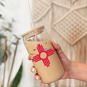 May include: A glass jar with a wooden lid and a red New Mexico state symbol on the side. The jar is filled with iced coffee and has a straw sticking out of the top.