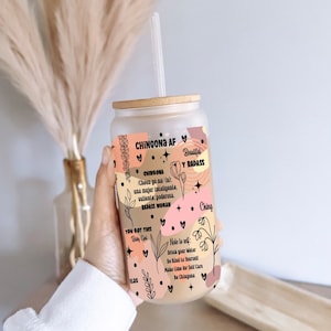 May include: A clear glass tumbler with a bamboo lid and a straw. The tumbler has a colourful design with the words "Chingona AF" and "Beautiful & Badass" in black text. The design also includes a floral pattern and the words "You got this" and "Baby Girl".