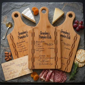 Könnte beinhalten: Drei hellbraune Schneidebretter aus Bambus mit Griffen, jeweils graviert mit dem Text Grandma's Pumpkin Rolls und einem handgeschriebenen Rezept. Die Bretter haben ein kariertes Randmuster und sind mit Käse, Crackern und Fleisch arrangiert.
