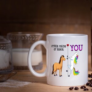 May include: White ceramic coffee mug with a unicorn and a horse graphic. The text on the mug reads "Other maids of honor" and "You" with a red heart.