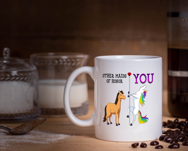 May include: White ceramic coffee mug with a unicorn and a horse graphic. The text on the mug reads "Other maids of honor" and "You" with a red heart.
