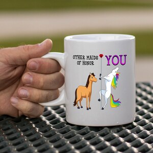 May include: White ceramic mug with a graphic of a unicorn and a horse. The unicorn is holding a pole with a red diamond at the top. The text on the mug reads "Other maids of honor" and "You".