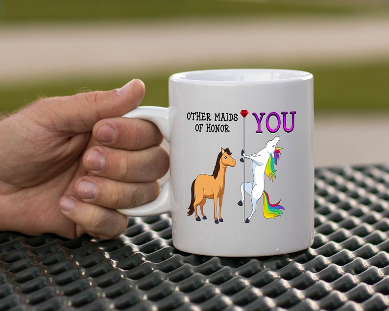 May include: White ceramic mug with a graphic of a unicorn and a horse. The unicorn is holding a pole with a red diamond at the top. The text on the mug reads "Other maids of honor" and "You".
