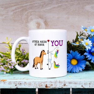 May include: White ceramic mug with a cartoon unicorn and horse. The mug reads "Other maids of honor YOU" with a red heart between the words.