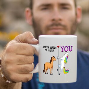 May include: White ceramic mug with a unicorn and a horse illustration. The text on the mug reads "Other maids of honor" and "You" with a red heart.