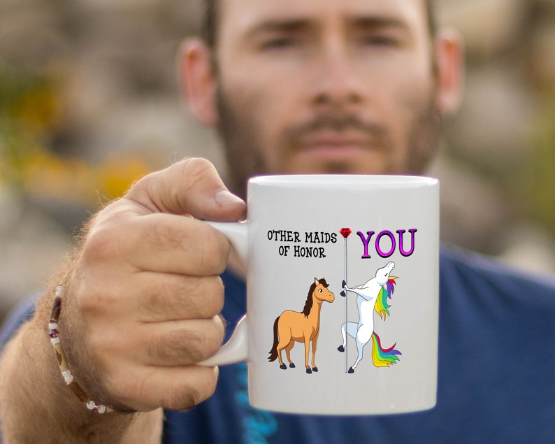 May include: White ceramic mug with a unicorn and a horse illustration. The text on the mug reads "Other maids of honor" and "You" with a red heart.