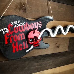 May include: A black electric guitar with a white snake and a red skull painted on it. The guitar has the words "Step Aside Cowboys From Hell" written on it in white and red paint.
