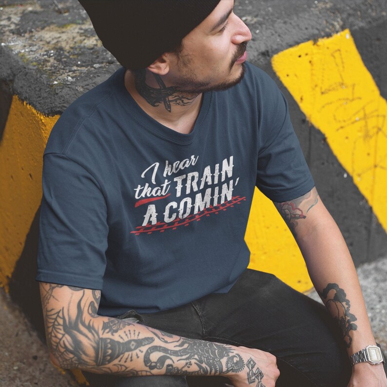 Puede incluir: Un hombre lleva una camiseta azul marino con el texto "I hear that TRAIN A COMIN'" en blanco con acentos rojos. La camiseta tiene un aspecto desgastado y el texto est&aacute; sobre un fondo de v&iacute;as de tren.