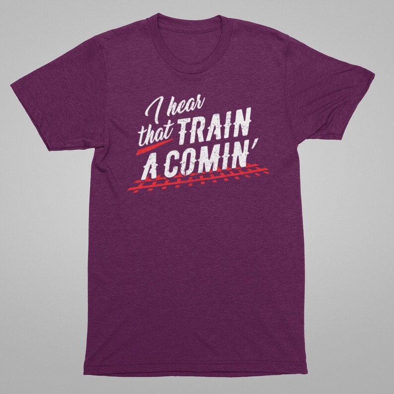 Puede incluir: Camiseta de manga corta de color morado con texto blanco que dice "I hear that TRAIN A COMIN'" con un gr&aacute;fico de v&iacute;a de tren roja debajo.