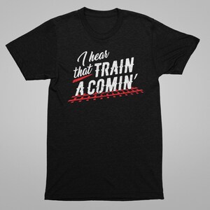 Puede incluir: Camiseta negra con texto blanco que dice "I hear that TRAIN A COMIN'" con v&iacute;as de tren rojas debajo.