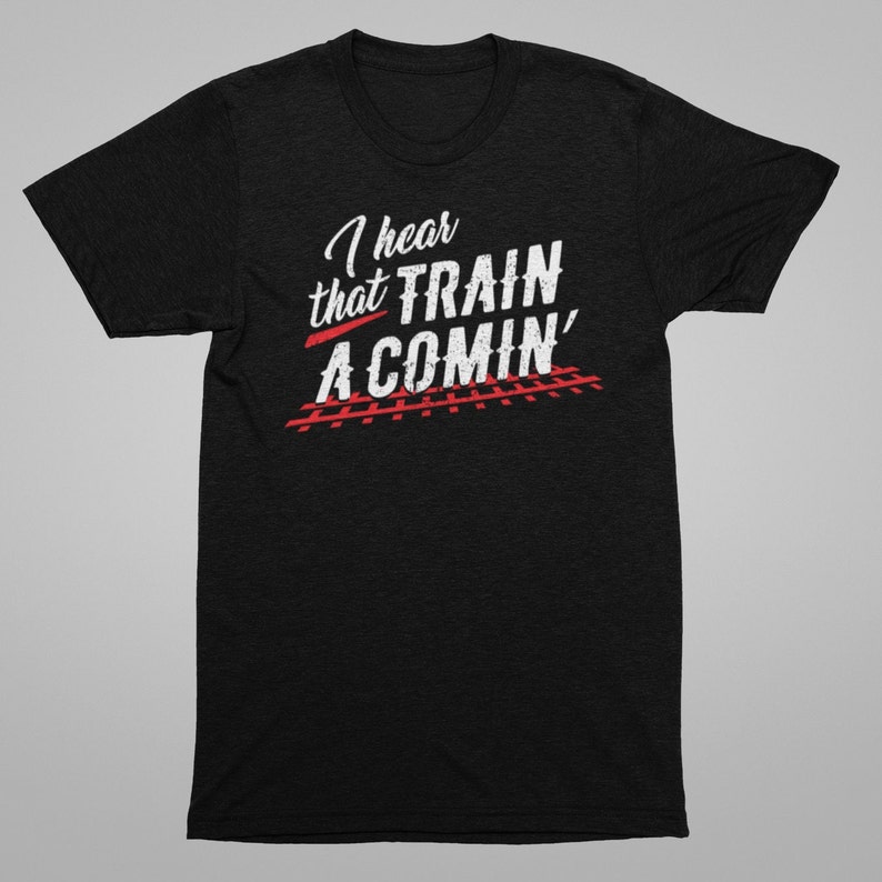 Puede incluir: Camiseta negra con texto blanco que dice "I hear that TRAIN A COMIN'" con v&iacute;as de tren rojas debajo.