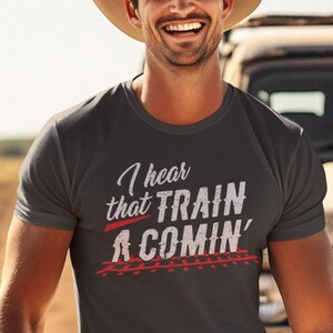Puede incluir: Una camiseta gris oscuro con un gr&aacute;fico blanco que dice "I hear that TRAIN A COMIN'" con una l&iacute;nea roja debajo de la palabra "TRAIN" y una imagen en blanco y negro de v&iacute;as de tren debajo del texto.
