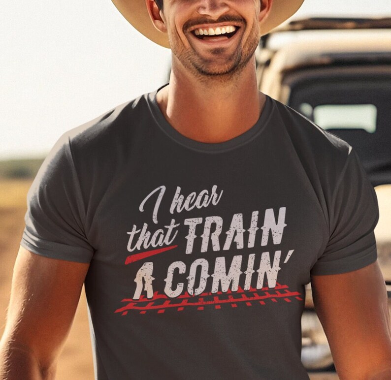 Puede incluir: Una camiseta gris oscuro con un gr&aacute;fico blanco que dice "I hear that TRAIN A COMIN'" con una l&iacute;nea roja debajo de la palabra "TRAIN" y una imagen en blanco y negro de v&iacute;as de tren debajo del texto.