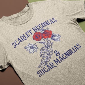May include: A heather gray t-shirt featuring the text "SCARLET BEGONIAS & SUGAR MAGNOLIAS" in blue. The graphic includes a skeleton hand holding red and blue flowers. The shirt is a casual wear item.