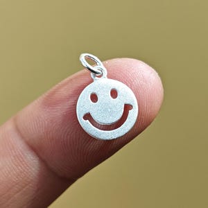 Puede incluir: Un pequeño dije de carita sonriente de color plateado con un lazo en la parte superior. El dije presenta un diseño clásico de carita sonriente con dos agujeros para los ojos y una boca curvada. El dije se sostiene entre dos dedos sobre un fondo borroso y marrón.