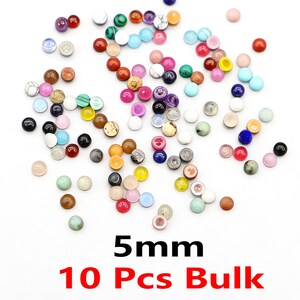 以下が含まれることがあります： 白い表面に散らばった、5mmのマルチカラーの丸いビーズのコレクション。ビーズは赤、青、緑、白など様々な色があります。下部のテキストは「5mm 10 Pcs Bulk」と表示されています。