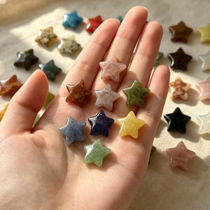 Puede incluir: Una colección de pequeñas piedras pulidas con forma de estrella en varios colores, incluyendo verde, azul, amarillo, rosa, marrón, negro y blanco. Varias de estas tallas lisas de cinco puntas se sostienen en una palma, otras están dispersas.