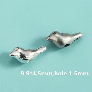 May include: Two silver-colored bird-shaped beads on a light blue background. The beads are detailed with small eyes and tail feathers. Text on the image indicates the beads are 9.9mm x 4.5mm with a 1.5mm hole.