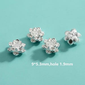 Puede incluir: Cuatro cuentas plateadas con forma de flor de loto, con un diseño detallado. Cada cuenta mide 9 mm x 5,3 mm, con un agujero de 1,9 mm. Se muestran sobre un fondo verde azulado claro.