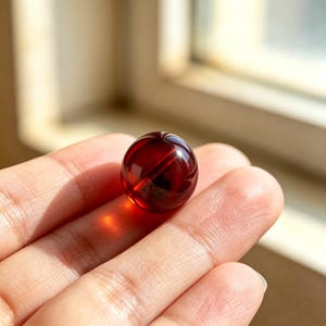 Puede incluir: Una pequeña cuenta esférica de color rojo oscuro translúcido. La superficie de la cuenta es lisa y pulida, reflejando la luz para crear un brillo intenso en su exterior redondeado, mostrando un tono carmesí rico y uniforme en toda ella.