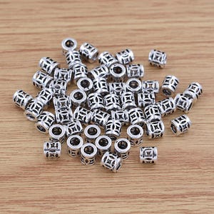 May include: A collection of silver-colored metal beads with a cylindrical shape and intricate openwork designs. The beads are scattered on a light brown wooden surface, showcasing their detailed patterns and reflective quality. These beads are suitable for jewelry making.