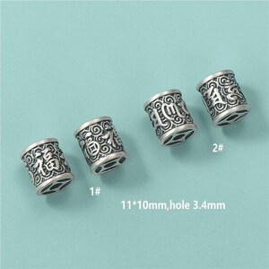 Peut inclure: Quatre perles cylindriques argentées avec des motifs gravés et des caractères chinois. Chaque perle mesure environ 11 mm x 10 mm, avec un trou de 3,4 mm. Présentées sur un fond bleu clair.