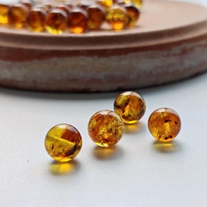 Peut inclure: Une collection de petites perles d'ambre sphériques aux teintes ambrées et translucides. Les perles présentent des inclusions internes naturelles et des variations de couleur, avec quatre perles individuelles au premier plan et d'autres derrière.
