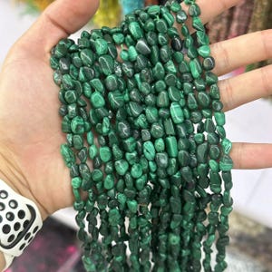 Pode incluir: Fios de contas de pedra malaquite polida em vários tons de verde. As contas são de formato e tamanho irregulares, com um acabamento liso e brilhante. A malaquite apresenta padrões e texturas naturais.