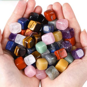 Puede incluir: Una colección de cubos de piedras preciosas pulidas y coloridas en varios colores, incluyendo rojo, azul, amarillo, rosa y verde. Las piedras preciosas se sostienen en manos ahuecadas, mostrando sus superficies lisas y variaciones naturales. Las piedras son de varios tamaños.