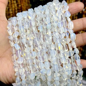 Puede incluir: Cadenas de cuentas de piedra preciosa de opalita pulida, de color blanquecino. Las cuentas tienen forma irregular y una calidad translúcida, con sutiles destellos iridiscentes. Las cuentas se sostienen en una mano, mostrando su belleza natural.
