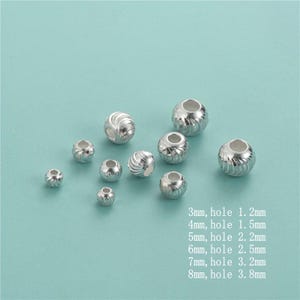 Puede incluir: Un conjunto de cuentas plateadas con un patrón en espiral, de varios tamaños. El texto indica los tamaños de las cuentas de 3 mm a 8 mm, con los tamaños de los agujeros. Estas cuentas son adecuadas para la fabricación de joyas y manualidades.