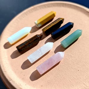 May include: Eight polished crystal points in various colors including yellow, brown, black, white, green, and pink. Each stone is shaped into a hexagonal prism with a pointed tip, arranged in a scattered pattern on a circular terracotta surface.