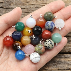 Puede incluir: Una colección de colgantes de piedras preciosas en varios colores, incluyendo verde, blanco, amarillo y azul. Cada colgante es una esfera con un lazo plateado para sujetar a una cadena. Las piedras tienen diferentes patrones y texturas.