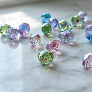 Puede incluir: Una colección de cuentas de vidrio facetado de diversas formas y tamaños, con un acabado iridiscente. Las cuentas muestran un espectro de colores que incluye verde claro, violeta suave y azul brillante, con superficies reflectantes.