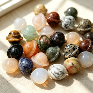 May include: A collection of small, polished gemstone carvings shaped like planets with rings. The stones feature various colours and patterns, including tiger's eye, dalmatian jasper, unakite, sodalite, white howlite, and translucent quartz varieties.
