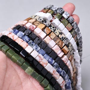 Puede incluir: Una mano muestra hilos de cuentas cuadradas de piedras preciosas en varios colores y patrones. Las cuentas incluyen diseños verdes, negros, blancos y moteados, ideales para la creación de joyas. Las cuentas están ensartadas, mostrando su forma uniforme y superficies pulidas.