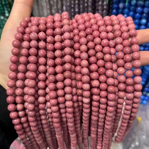 Könnte beinhalten: Stränge aus rosa Edelsteinperlen. Die Perlen sind rund und haben eine glatte, polierte Oberfläche. Die Perlen haben eine staubrosa Farbe und scheinen aufgefädelt zu sein. Der Hintergrund ist unscharf, aber andere Farben von Perlen sind sichtbar.
