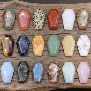 May include: A collection of coffin-shaped gemstone pendants in various colors and patterns. The gemstones include red, white, black-spotted, and tiger's eye, arranged on a wooden surface. These pendants are ideal for jewelry making.