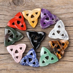 Op de afbeelding: Een verzameling kleurrijke, gepolijste stenen Keltische knoop hangers. De hangers zijn in verschillende kleuren, waaronder rood, geel, paars, groen, zwart, wit, bruin, roze, blauw en groen. Elke hanger heeft een driehoekige vorm.