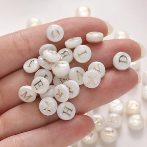 Op de afbeelding: Een verzameling witte, ronde kralen met gouden letters, die verschillende letters van het alfabet spellen. De kralen worden in een hand gehouden, wat hun grootte en ontwerp laat zien. De kralen hebben een parelmoerglans.