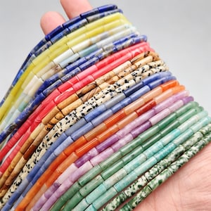 May include: A hand holds multiple strands of colorful, rectangular beads. The beads are in various colors, including blue, yellow, red, brown, and green. The beads are made of stone and are strung together, ready for jewelry making.
