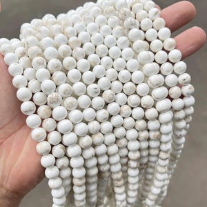 May include: Strands of white gemstone beads with subtle brown marbling. The round beads are uniform in size and shape, creating a smooth, consistent texture. These beads are ideal for jewelry making, such as necklaces and bracelets.